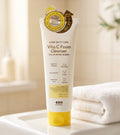 A tube of BearaGo Dark Spot Care Vita C Foam Cleanser stands on a white tray next to a folded white towel in a softly lit bathroom setting.
