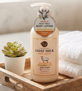 A bottle of Darlie's Pure & Natural Goat Milk Body Lotion with Manuka Honey Ointment, styled on a wooden tray next to a small potted succulent and a stack of white towels.