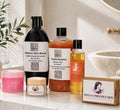 A collection of Paula's Precious Skin and By Paula's body care products arranged on a white marble countertop in a bathroom setting. The products include a black bottle of Radiant Skin Boost Lightening Body Lotion, a Triple-Action Shower Gel bottle filled with orange beads, a yellow bottle of Face Brightening Shampoo, a pink jar of Deep Cleansing Milk, a small cream jar with a wooden lid, and a wooden box of Face Brightening Shampoo.