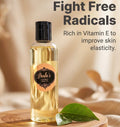 A bottle of Paula's Vitamins Body Oil with a black cap and a peach-colored label, displayed on a round wooden tray with green leaves. The background is a warm, softly blurred beige setting with text overlays reading 'Fight Free Radicals' and 'Rich in Vitamin E to improve skin elasticity.