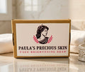 A rectangular cardboard box of Paula's Precious Skin Face Brightening Soap, featuring a white label with a black classical profile illustration and pink laurel leaves, set on a marble surface with white towels in the background.