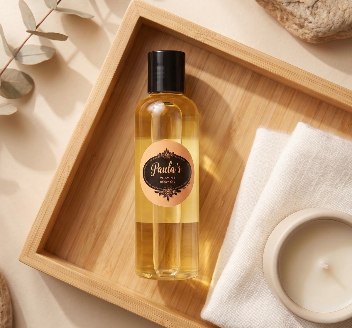 A bottle of Paula's Handmade Body Oil with a black cap and a peach label, displayed on a wooden tray next to a folded white towel and a candle.