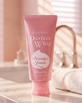 A pink squeeze tube of Senka Perfect Whip Beauty Foam Collagen in foam, standing on a marble countertop with a blurred bathroom sink and faucet in the background.