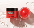 Brightening Lip Balm by By: Paula's in a coral-red jar with a black lid, shown next to an open jar of the same balm on a white background with decorative water droplets.