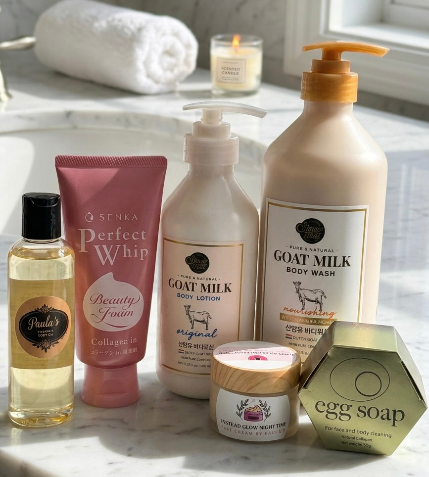 A collection of personal care products arranged on a white marble surface near a window: a bottle of Puja's Perfect Whip collagen-in enriched liquid, a pink tube of Senka Beauty Potion, a pump bottle of Pure Encapsulations Goat Milk Body Lotion, a large pump bottle of Pure Encapsulations Goat Milk Body Wash, a small jar of Roman Glow Night Cream, and a hexagonal box of egg soap.