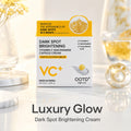 A box of OOTD+ VC+ Dark Spot Brightening Vitamin C Niacinamide Capsule Cream is displayed on a reflective white surface with decorative glass vases and warm lighting in the background. The packaging is white with yellow and orange accents, and the text 'Luxury Glow Dark Spot Brightening Cream' is written below.